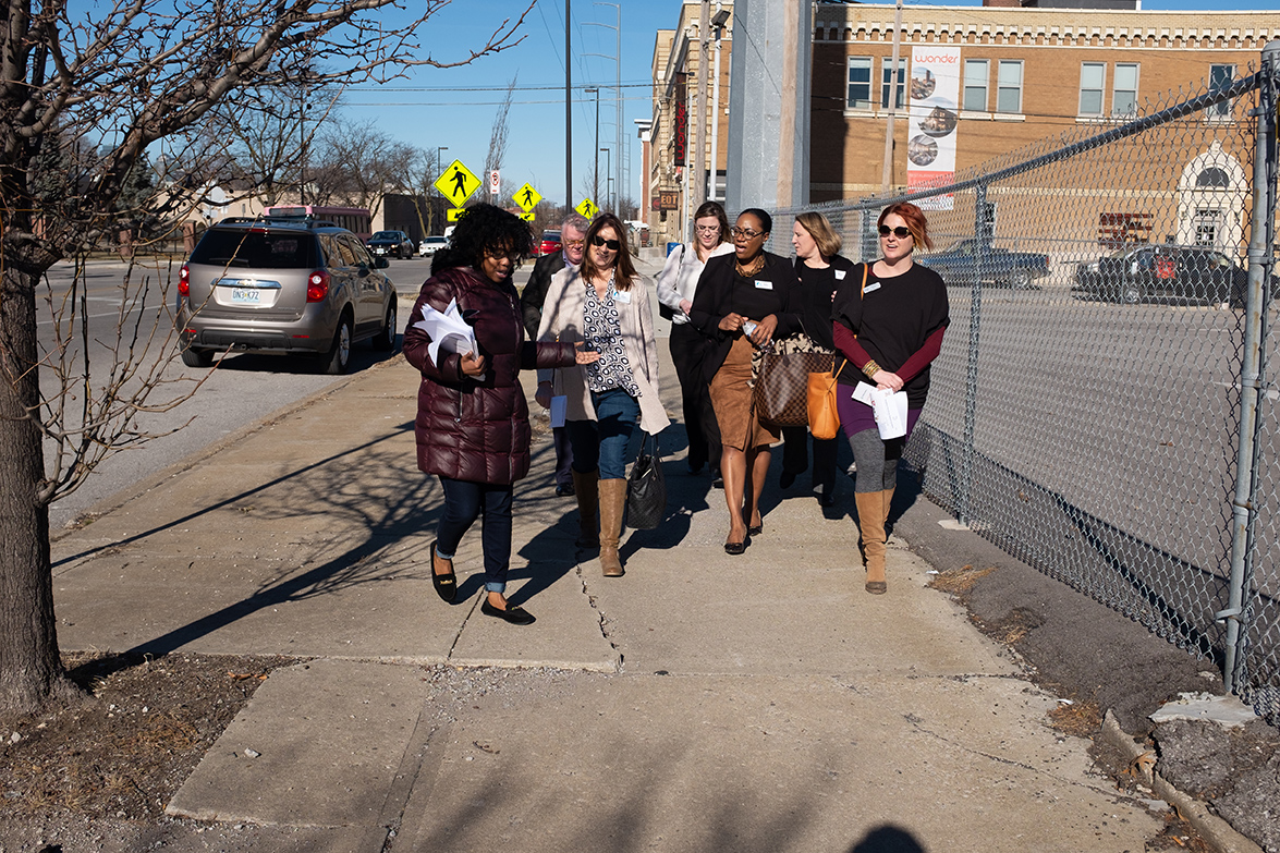 A group of women of mixed ages and races are walking and talking together on a wide, cracked sidewalk.