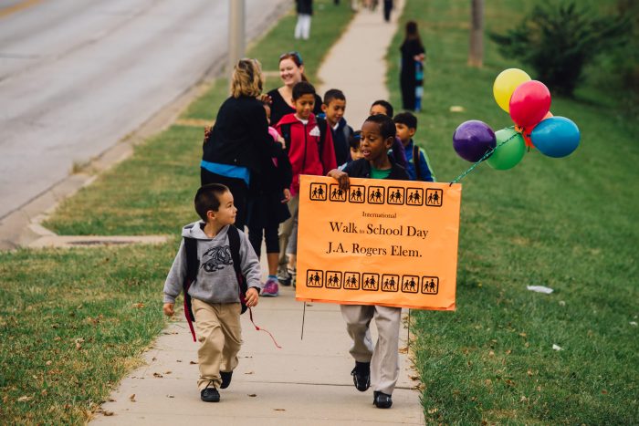 Students walking to J.A. Rogers Elementary in 2015. They hold a sign and balloons celebrating Walk to School Day.