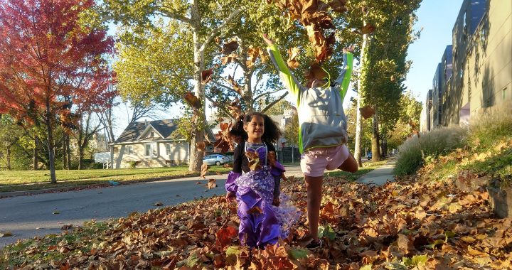 Two young girls frolic in a pile of orange and yellow leaves on the sidewalk. One is wearing a purple and black witch costume, and one is wearing athletic gear.