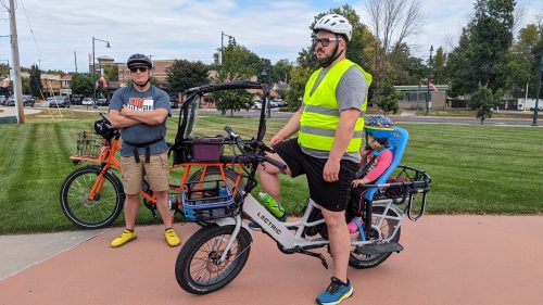 Two parents pose with their electric bikes. Both bikes have special carriers for children who are too young to ride their own bike. 