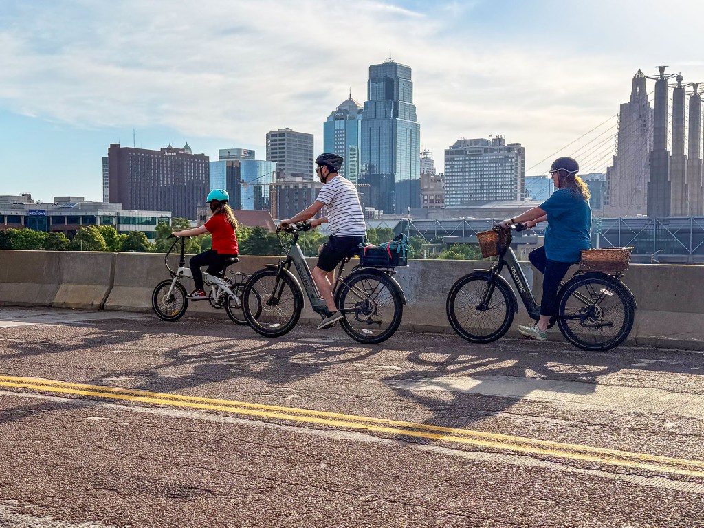 Two adults and a child ride e-bikes on the street. The Kansas City skyline rises in the distance.