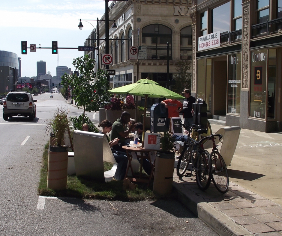 People are relaxing together and enjoying a car-parking spot that has been converted into a parklet, complete with a big shade umbrella, live plants, real sheets of sod, chairs, and small tables.