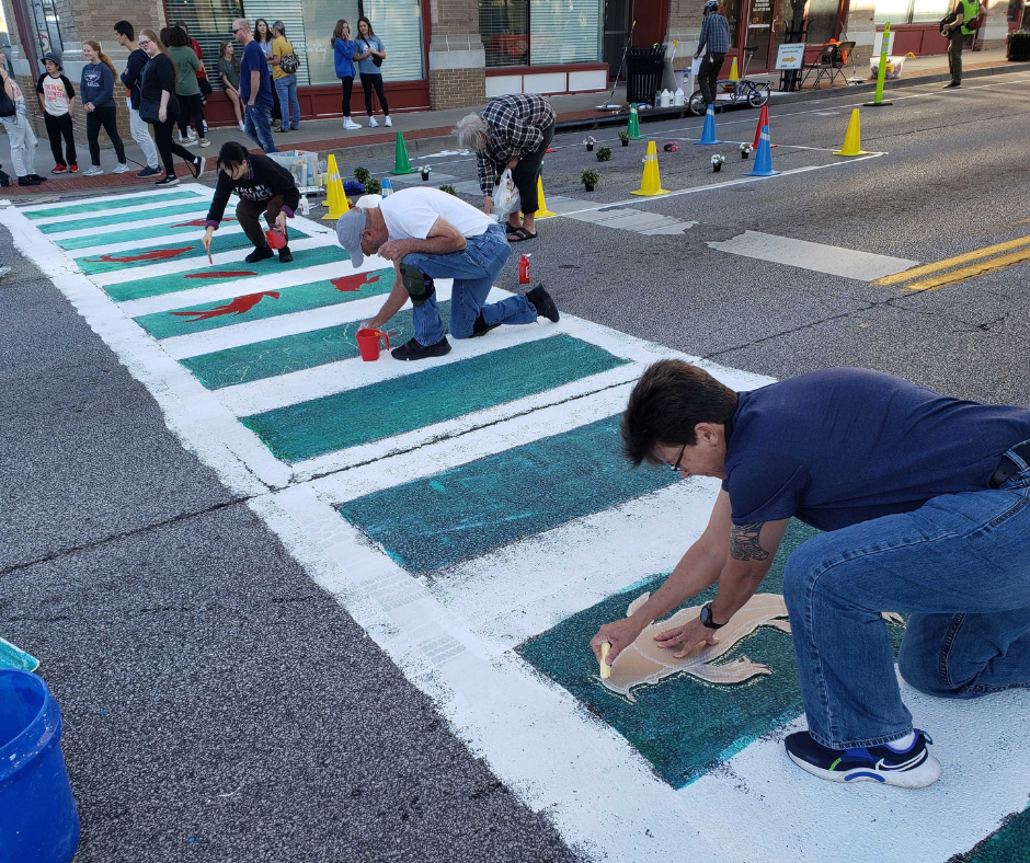 Volunteers paint a colorful crosswalk in a lively downtown area. The crosswalk is white and green with brightly colored fish.